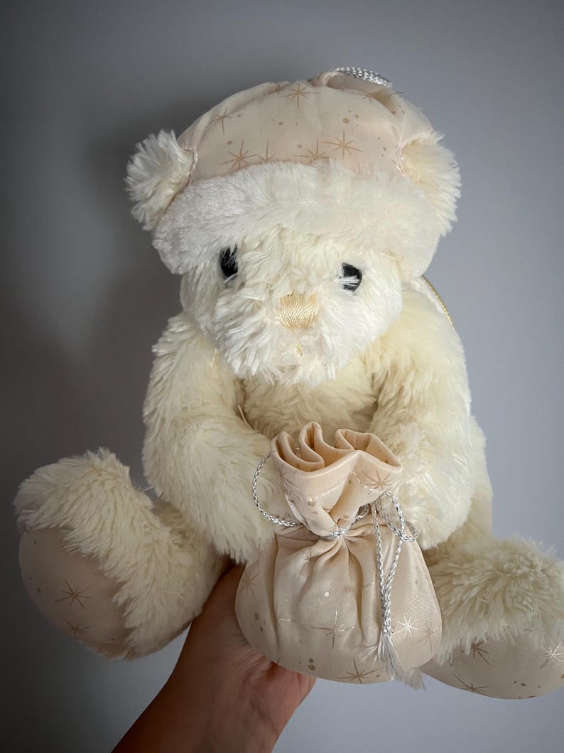 May include: A cream-colored teddy bear wearing a matching hat with gold starburst patterns. The plush bear holds a small gift bag with a silver drawstring. The bear's paws and hat feature the same starburst design.