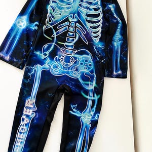 May include: A black jumpsuit with a glowing blue skeleton design. The skeleton is outlined in white, with blue lightning bolt accents. The jumpsuit has long sleeves and legs, ideal for Halloween or fancy dress.