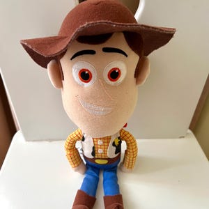 Puede incluir: Un peluche de un personaje de dibujos animados de vaquero. El juguete tiene un sombrero marr&oacute;n, pelo casta&ntilde;o y una cara sonriente. Viste una camisa a cuadros amarilla, un chaleco blanco, pantalones azules y botas marrones. El juguete est&aacute; sentado.