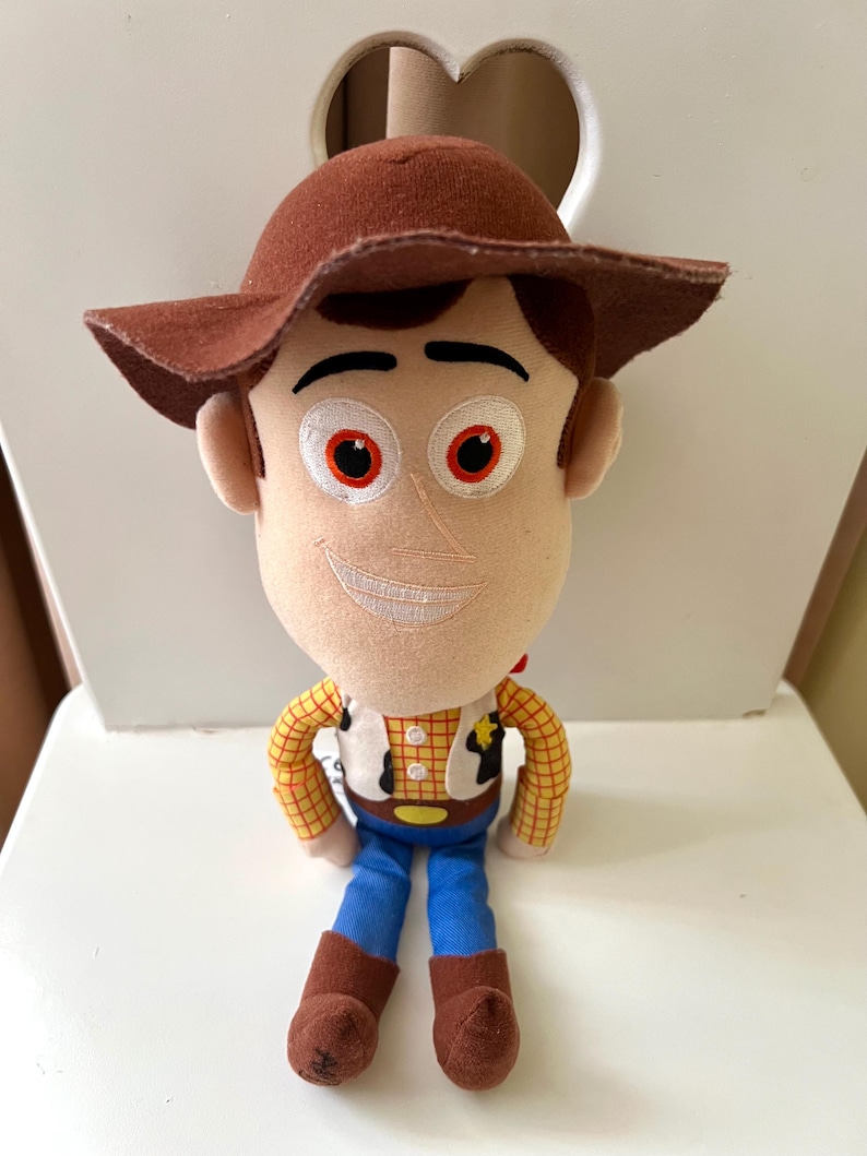 Puede incluir: Un peluche de un personaje de dibujos animados de vaquero. El juguete tiene un sombrero marr&oacute;n, pelo casta&ntilde;o y una cara sonriente. Viste una camisa a cuadros amarilla, un chaleco blanco, pantalones azules y botas marrones. El juguete est&aacute; sentado.