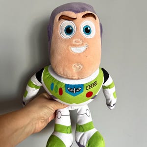 May include: A plush Buzz Lightyear doll from the Toy Story franchise. The doll features a light peach face with blue eyes, a purple head, and a green, white, and black spacesuit. The doll has the word "Lightyear" on the chest.