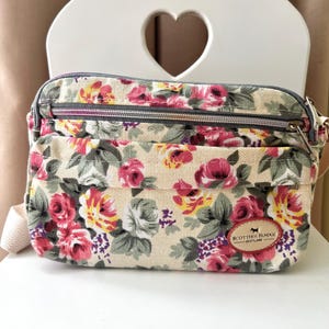 Op de afbeelding: Een bloemen crossbody tas met een beige achtergrond en een patroon van roze, rode, gele en witte rozen. De tas heeft een grijze rits en een kleine patch met de tekst "Scottish Renzos Scotland". De tas staat op een witte stoel.