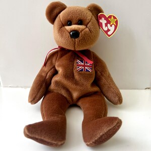 May include: A brown teddy bear plush toy with a black nose, a red ribbon, and a small Union Jack flag patch. The bear has a Ty heart-shaped tag attached. The plush is sitting on a white surface.