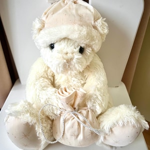May include: A cream-colored teddy bear wearing a matching hat and holding a small bag. The hat and bag feature a star pattern. The bear is sitting on a white chair, creating a cozy and whimsical scene.