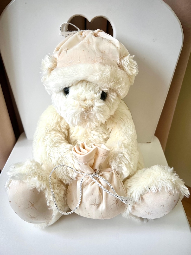 May include: A cream-colored teddy bear wearing a matching hat and holding a small bag. The hat and bag feature a star pattern. The bear is sitting on a white chair, creating a cozy and whimsical scene.