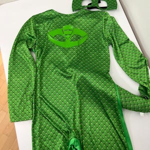 Könnte beinhalten: Grünes Kostüm mit Reptilienschuppenmuster und passender Maske. Der langärmlige Overall hat ein großes Emblem auf dem Rücken und einen Schwanz. Die Maske hat Augenausschnitte und eine spitze Oberseite. Das Kostüm ist für Kinder.
