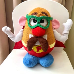 Può includere: Un giocattolo di peluche a forma di patata con un tema da supereroe. Il giocattolo ha un corpo marrone, un naso rosso e dei baffi marroni. Indossa occhiali verdi, un mantello rosso e pantaloni blu. Il petto ha un emblema giallo con la lettera "P".