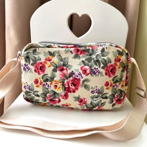 Op de afbeelding: Crossbody tas met bloemenprint op een cr&egrave;mekleurige achtergrond, met rode, roze en gele rozen en groene bladeren. De tas heeft een verstelbare cr&egrave;mekleurige riem en een zilveren rits. De tas staat op een witte stoel.