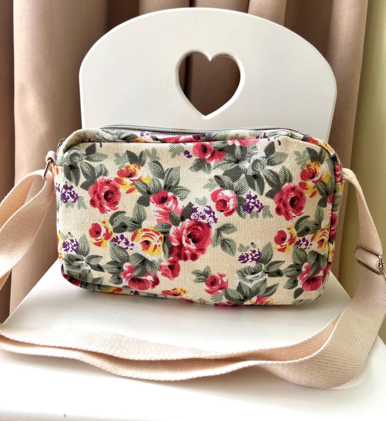 Op de afbeelding: Crossbody tas met bloemenprint op een cr&egrave;mekleurige achtergrond, met rode, roze en gele rozen en groene bladeren. De tas heeft een verstelbare cr&egrave;mekleurige riem en een zilveren rits. De tas staat op een witte stoel.