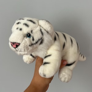 Peut inclure: Une peluche tigre blanc avec une fourrure douce à rayures noires, un nez rose et des yeux bleu vif. L'animal en peluche est représenté dans une posture quadrupède avec la tête légèrement tournée sur le côté, mettant en valeur ses détails.