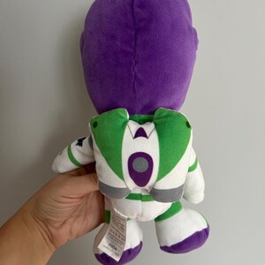 Pode incluir: Um boneco de pel&uacute;cia Buzz Lightyear, visto por tr&aacute;s, apresenta uma cabe&ccedil;a roxa e um design de roupa espacial branca e verde. O boneco tem p&eacute;s brancos com detalhes roxos e asas verdes. O boneco &eacute; segurado por uma m&atilde;o contra um fundo liso.