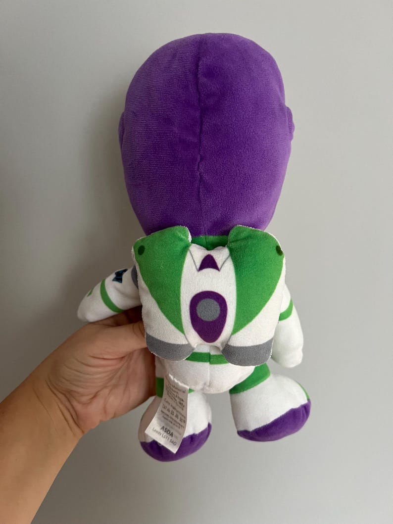 Pode incluir: Um boneco de pel&uacute;cia Buzz Lightyear, visto por tr&aacute;s, apresenta uma cabe&ccedil;a roxa e um design de roupa espacial branca e verde. O boneco tem p&eacute;s brancos com detalhes roxos e asas verdes. O boneco &eacute; segurado por uma m&atilde;o contra um fundo liso.