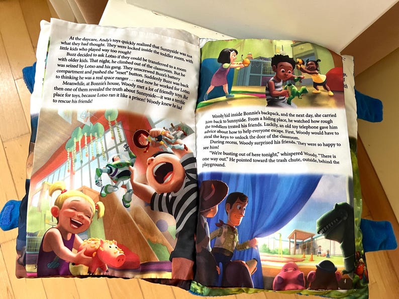 May include: A soft fabric book featuring Toy Story characters with printed text on two open pages. The left page shows a child playing with a pink piggy bank toy while Buzz Lightyear is in the background. The right page shows Woody and Rex.
