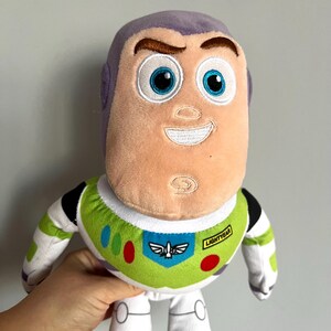 May include: A plush Buzz Lightyear doll from the Toy Story franchise. The doll features a light purple head, a peach-colored face, and a green and white spacesuit. The spacesuit has colorful accents and the word "Lightyear" is printed on the chest.