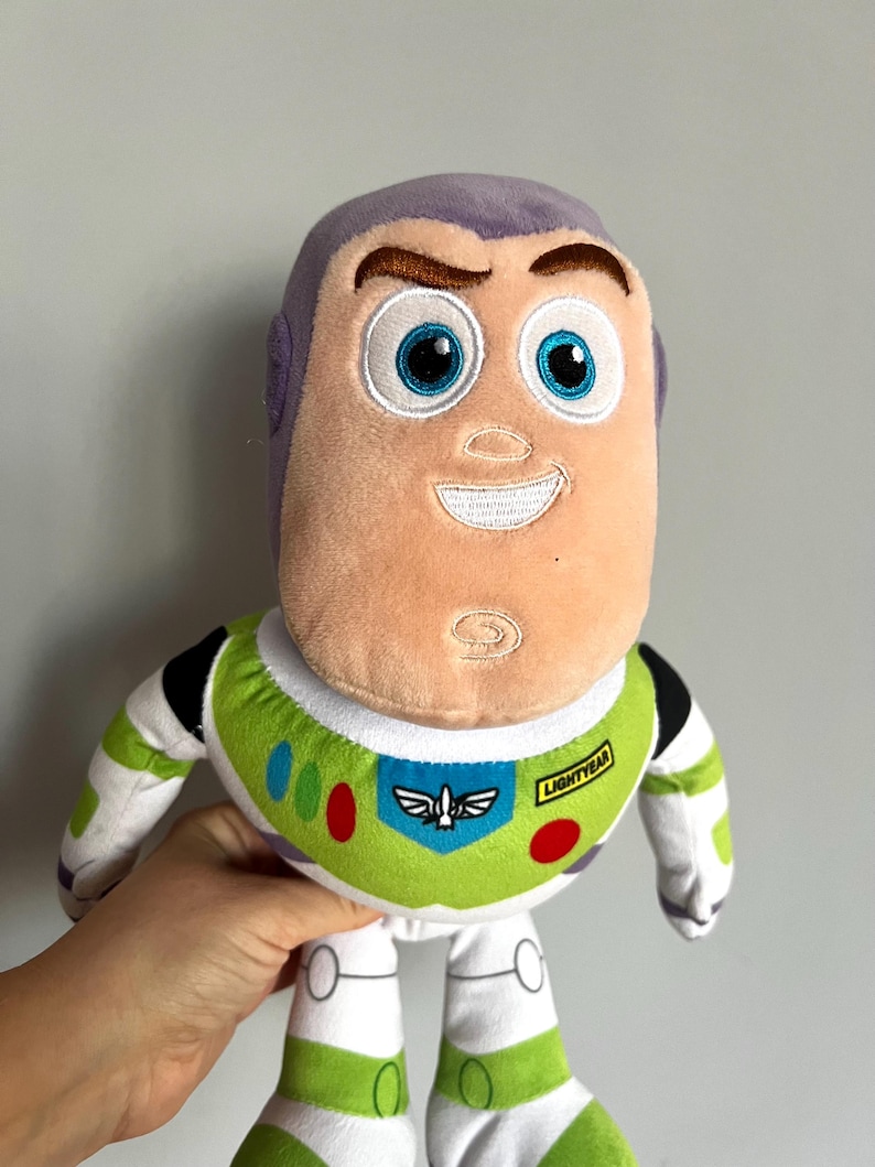 May include: A plush Buzz Lightyear doll from the Toy Story franchise. The doll features a light purple head, a peach-colored face, and a green and white spacesuit. The spacesuit has colorful accents and the word "Lightyear" is printed on the chest.