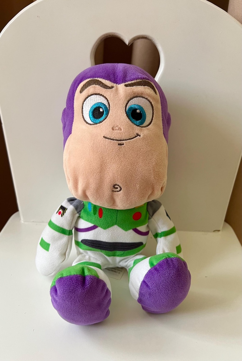 Pode incluir: Um brinquedo de pel&uacute;cia de Buzz Lightyear, um personagem da franquia Toy Story. O brinquedo tem cabe&ccedil;a e p&eacute;s roxos, rosto bronzeado claro e um traje espacial verde e branco. O brinquedo est&aacute; sentado em uma cadeira branca.