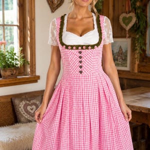 May include: A pink and white gingham dirndl dress featuring a white blouse with short lace sleeves, a square neckline, a dark green velvet trim adorned with small pink floral accents, and a row of heart-shaped buttons down the front bodice.