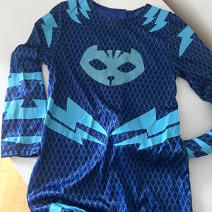 May include: A dark blue costume with a diamond pattern and light blue lightning bolt designs. The costume features a cat-like mask on the chest and lightning bolt accents on the sleeves and shoulders. The costume is likely for a child.