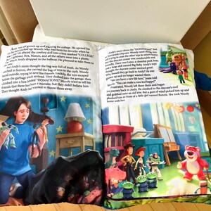 May include: A soft fabric book featuring Toy Story illustrations and printed text on two open pages. The left page shows a character holding toys, while the right page depicts a group of animated characters including a pink bear in a room setting.