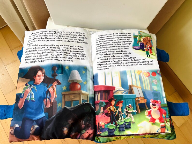 May include: A soft fabric book featuring Toy Story illustrations and printed text on two open pages. The left page shows a character holding toys, while the right page depicts a group of animated characters including a pink bear in a room setting.