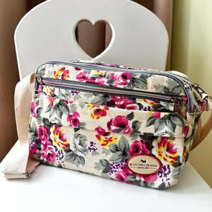 Op de afbeelding: Een crossbodytas met bloemenpatroon, met een cr&egrave;mekleurige achtergrond en een patroon van roze, gele en paarse rozen. De tas heeft een beige riem en een kleine bruine leren patch met tekst en een honden silhouet.