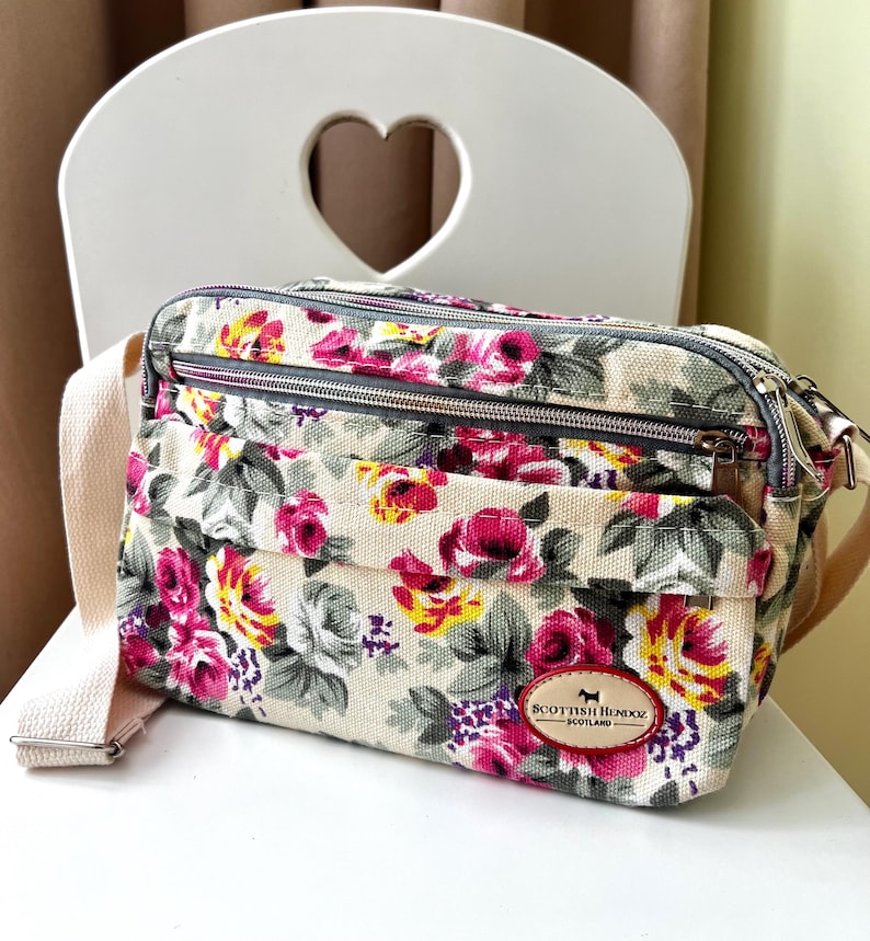 Op de afbeelding: Een crossbodytas met bloemenpatroon, met een cr&egrave;mekleurige achtergrond en een patroon van roze, gele en paarse rozen. De tas heeft een beige riem en een kleine bruine leren patch met tekst en een honden silhouet.