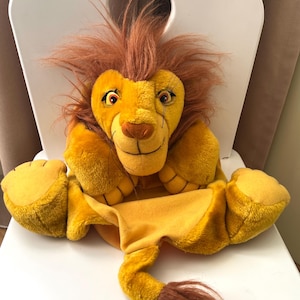 Puede incluir: Títere de león amarillo con melena y cola marrones. El títere tiene una cara sonriente con ojos detallados y una nariz marrón. El león está sentado en una silla blanca.