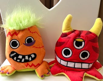 May include: Two plush monster toys. One is orange with green hair, black eyes, and a stitched mouth. The other is red with yellow horns, three black eyes, and a white stitched mouth. Both have a cartoonish design.