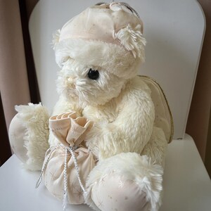 May include: A plush, cream-colored teddy bear with a matching hat and a small bag. The bear has fluffy fur, black eyes, and is seated on a white chair. The bag is tied with a silver string and has a star pattern.