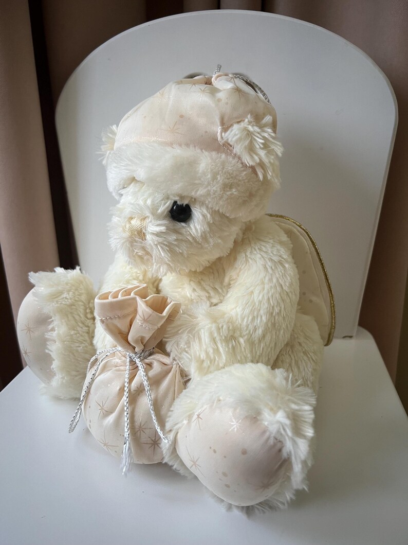 May include: A plush, cream-colored teddy bear with a matching hat and a small bag. The bear has fluffy fur, black eyes, and is seated on a white chair. The bag is tied with a silver string and has a star pattern.