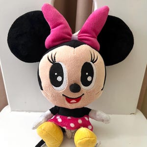 May include: A plush Minnie Mouse doll with black ears, a pink bow, and a pink and white polka dot dress. The doll has large black eyes, a red smile, and yellow shoes. The doll is sitting on a white chair.