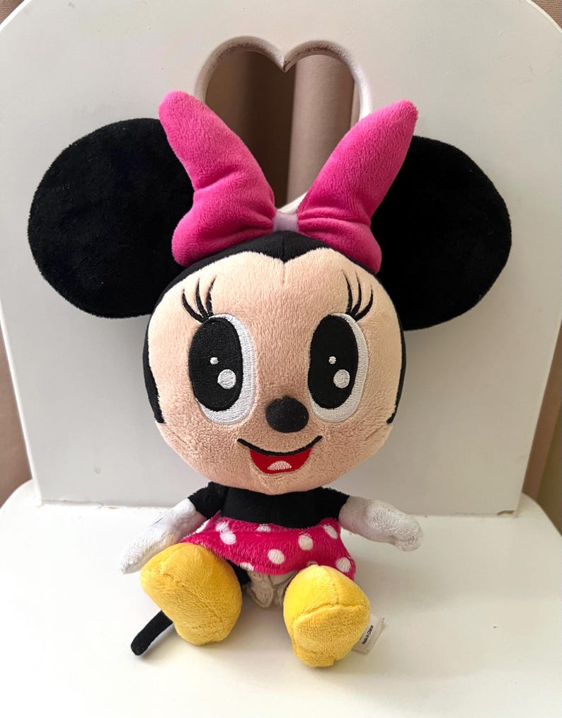 May include: A plush Minnie Mouse doll with black ears, a pink bow, and a pink and white polka dot dress. The doll has large black eyes, a red smile, and yellow shoes. The doll is sitting on a white chair.