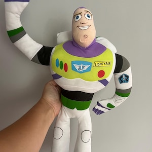 May include: A plush Buzz Lightyear doll from the Toy Story franchise. The doll features a white and green spacesuit with a purple collar and accents. The doll's arm is raised in a waving gesture. The word "LIGHTYEAR" is printed on the chest.