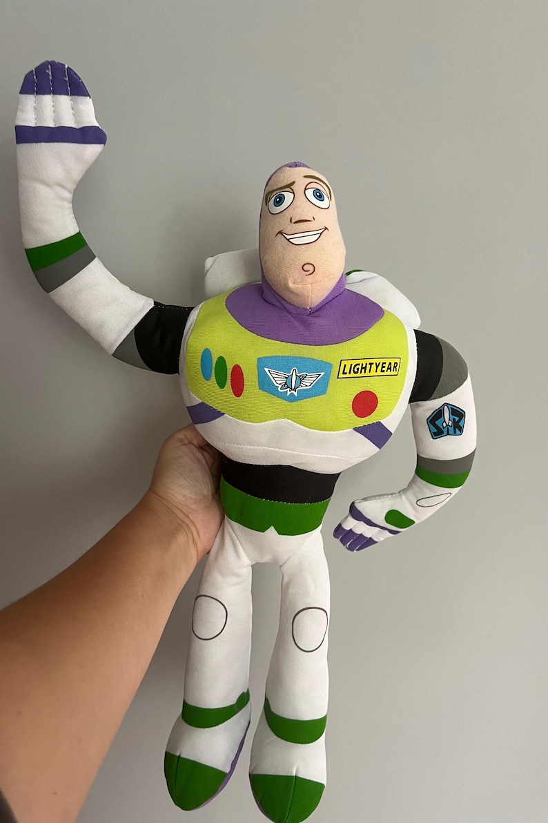 May include: A plush Buzz Lightyear doll from the Toy Story franchise. The doll features a white and green spacesuit with a purple collar and accents. The doll's arm is raised in a waving gesture. The word "LIGHTYEAR" is printed on the chest.