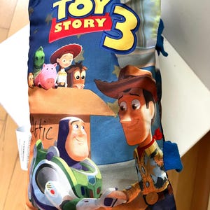 May include: A rectangular pillow featuring the Disney Pixar Toy Story 3 logo at the top. The graphic displays Buzz Lightyear and Woody shaking hands in the foreground, with Jessie, Bullseye, Rex, and Hamm visible above a box labeled Attic.