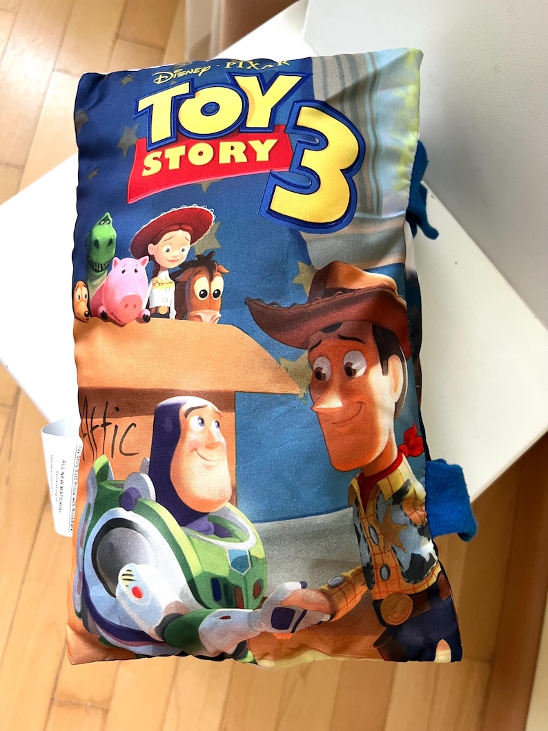 May include: A rectangular pillow featuring the Disney Pixar Toy Story 3 logo at the top. The graphic displays Buzz Lightyear and Woody shaking hands in the foreground, with Jessie, Bullseye, Rex, and Hamm visible above a box labeled Attic.