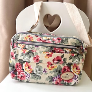 Op de afbeelding: Crossbody tas met bloemenprint op een cr&egrave;mekleurige achtergrond, met rode, roze en gele rozen, groene bladeren en paarse accenten. De tas heeft meerdere vakken met ritssluiting, een beige verstelbare riem en een label met "Scottish Henxx Scotland".
