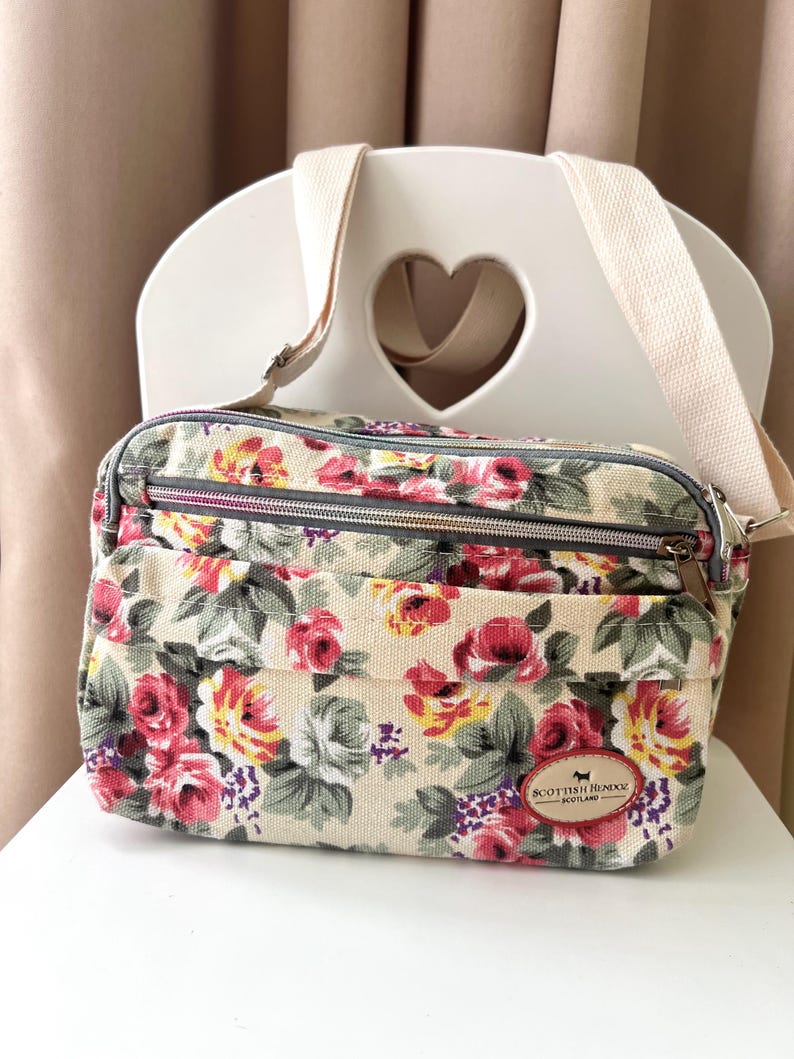 Op de afbeelding: Crossbody tas met bloemenprint op een cr&egrave;mekleurige achtergrond, met rode, roze en gele rozen, groene bladeren en paarse accenten. De tas heeft meerdere vakken met ritssluiting, een beige verstelbare riem en een label met "Scottish Henxx Scotland".