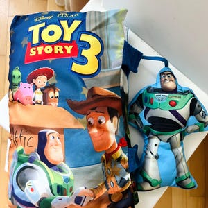 May include: A rectangular Toy Story 3 pillow featuring Woody, Buzz Lightyear, Jessie, Rex, Hamm, and Bullseye, accompanied by a separate, smaller, die-cut plush toy of Buzz Lightyear attached by a blue fabric strap to the side of the pillow.