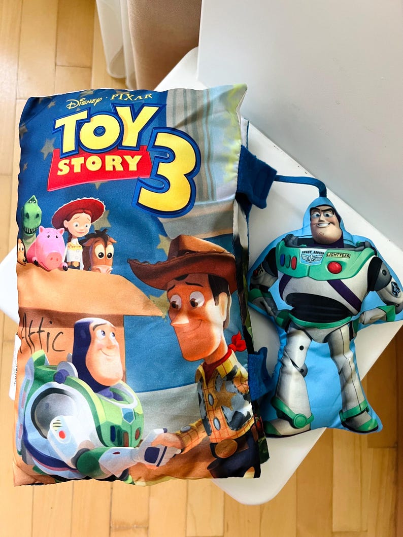 May include: A rectangular Toy Story 3 pillow featuring Woody, Buzz Lightyear, Jessie, Rex, Hamm, and Bullseye, accompanied by a separate, smaller, die-cut plush toy of Buzz Lightyear attached by a blue fabric strap to the side of the pillow.