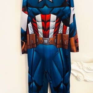 May include: Blue Captain America costume with a white star on the chest and a brown belt. The long-sleeved jumpsuit features red and white accents and the letter "A" on the sleeve. A superhero-themed outfit.
