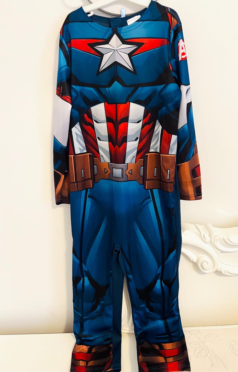 May include: Blue Captain America costume with a white star on the chest and a brown belt. The long-sleeved jumpsuit features red and white accents and the letter "A" on the sleeve. A superhero-themed outfit.