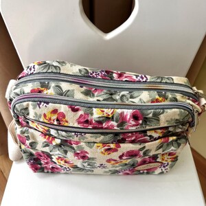 Op de afbeelding: Crossbody tas met bloemenprint en meerdere compartimenten met ritssluiting. De tas heeft een cr&egrave;mekleurige achtergrond met een patroon van roze, rode en gele bloemen en groene bladeren. De ritsen zijn grijs en de tas heeft een cr&egrave;mekleurige riem.