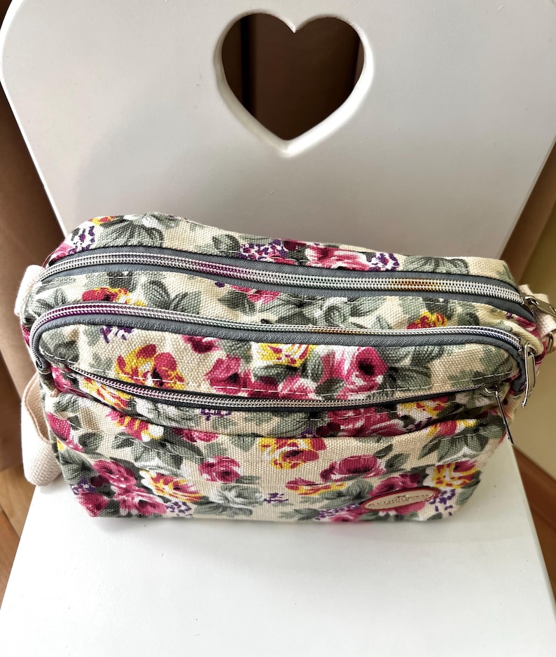 Op de afbeelding: Crossbody tas met bloemenprint en meerdere compartimenten met ritssluiting. De tas heeft een cr&egrave;mekleurige achtergrond met een patroon van roze, rode en gele bloemen en groene bladeren. De ritsen zijn grijs en de tas heeft een cr&egrave;mekleurige riem.