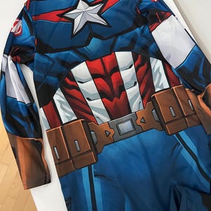 May include: A Captain America costume featuring a blue, red, and white design. The long-sleeved outfit includes a star emblem on the chest and a brown belt. The costume is designed to resemble the superhero's uniform.