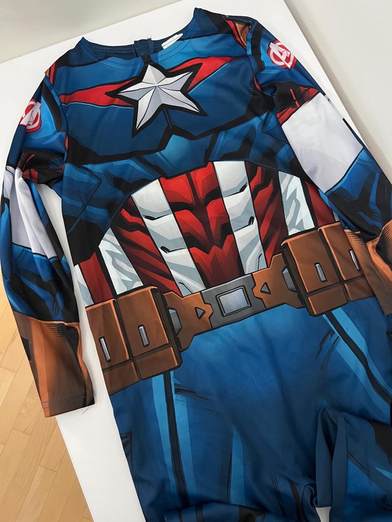 May include: A Captain America costume featuring a blue, red, and white design. The long-sleeved outfit includes a star emblem on the chest and a brown belt. The costume is designed to resemble the superhero's uniform.