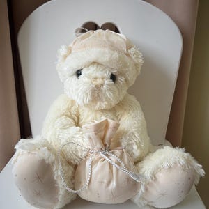 May include: A cream-colored teddy bear wearing a matching hat, seated on a white chair. The bear holds a small, light pink pouch with a silver drawstring. The pouch and hat feature a subtle star pattern. The bear's paws also have the star pattern.