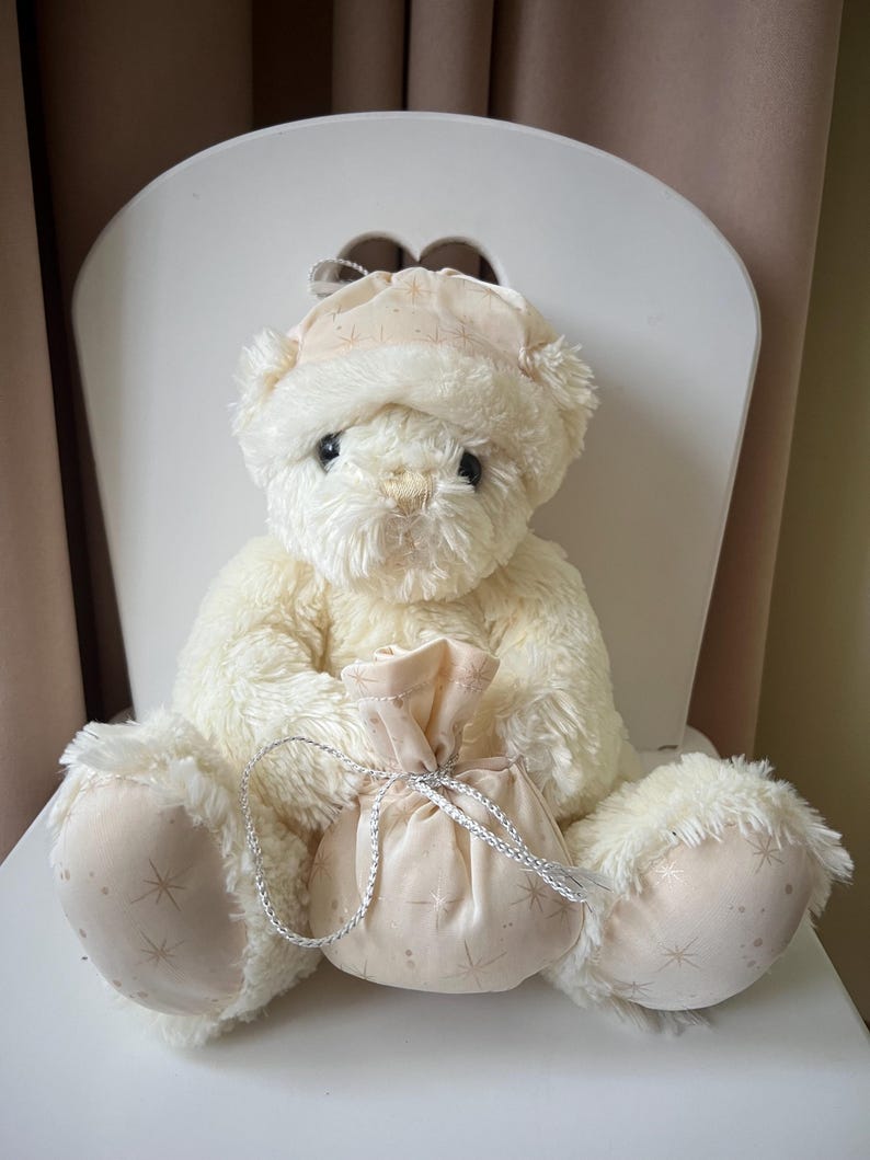 May include: A cream-colored teddy bear wearing a matching hat, seated on a white chair. The bear holds a small, light pink pouch with a silver drawstring. The pouch and hat feature a subtle star pattern. The bear's paws also have the star pattern.