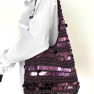 May include: A dark purple shoulder bag with large, round sequins. The bag is crocheted with a beaded trim and a long strap. The sequins are arranged in horizontal rows, creating a shimmering effect. The bag is displayed on a mannequin.
