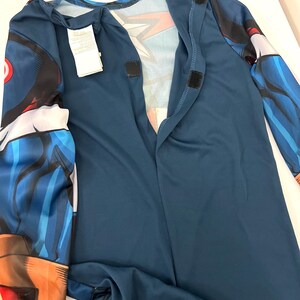 May include: A blue superhero costume with long sleeves and a mock neck. The costume has a graphic design on the sleeves and chest, with hook and loop fasteners down the front. A white tag is attached to the costume.
